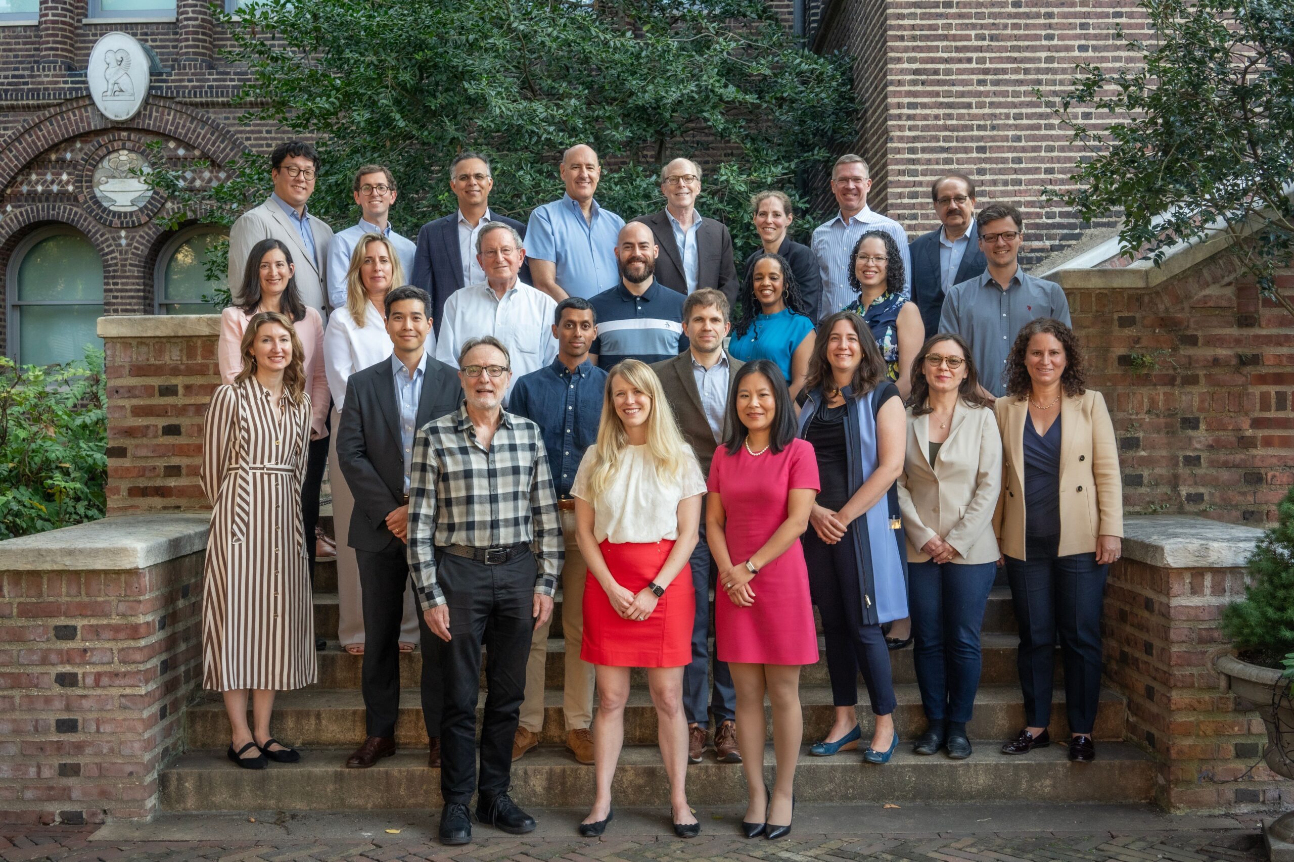 Faculty-Group-Picture-2024 A group of people, dressed in business-casual attire, are posing on steps in front of a brick building with greenery.